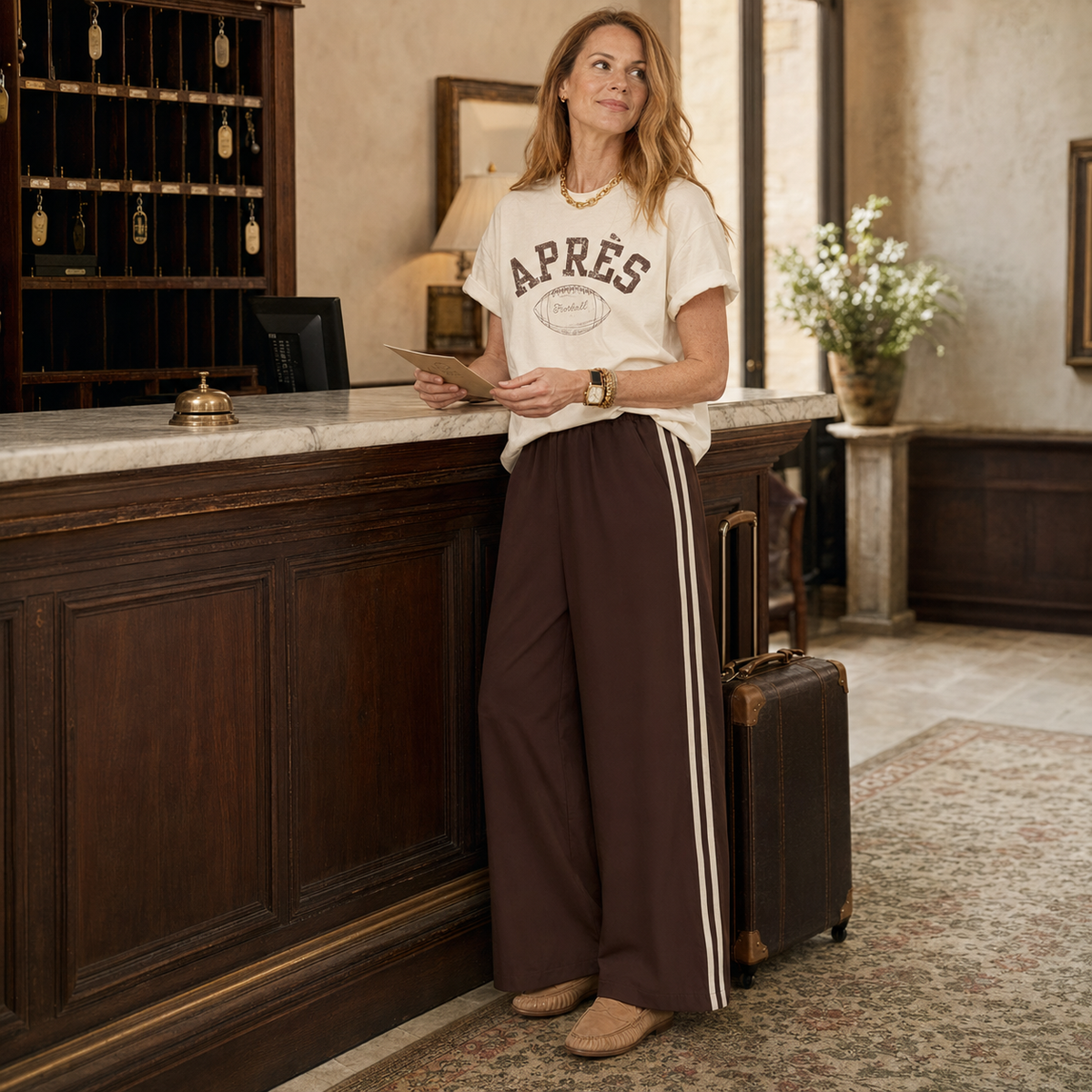 A woman stands in a hotel lobby next to a wooden reception desk, wearing a cream graphic t-shirt that says 'APRÈS' and dark brown wide-leg track pants with white side stripes. She is holding a tablet, with a brown suitcase beside her.