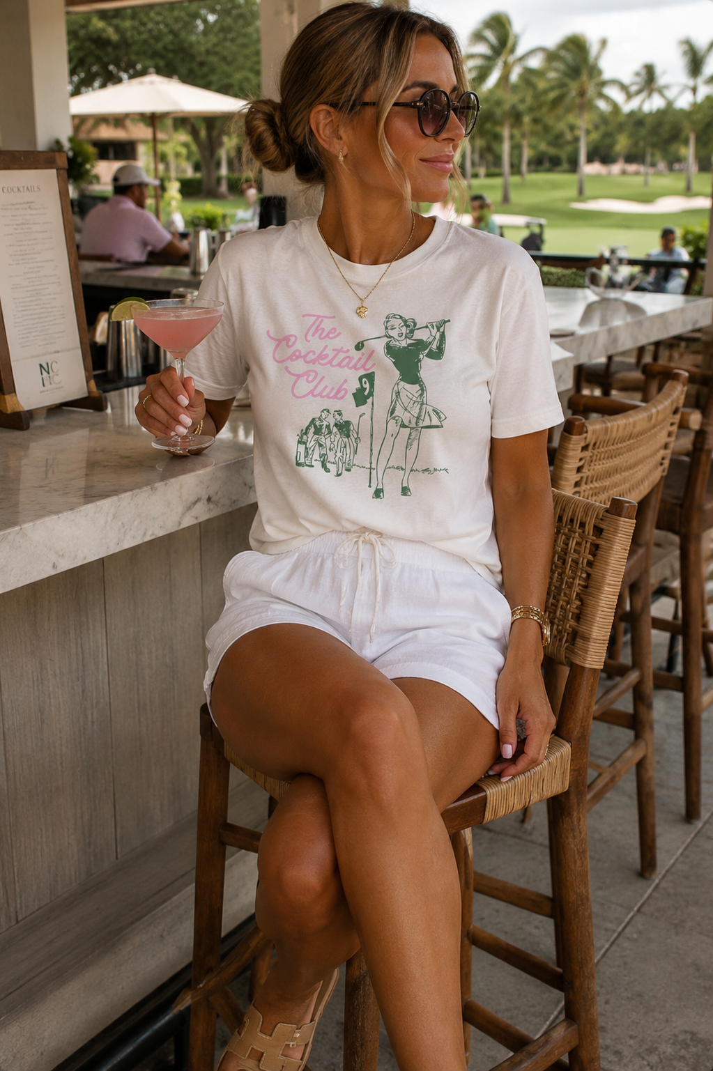 Woman sitting at a bar holding a pink drink, wearing a white t-shirt with a graphic design.