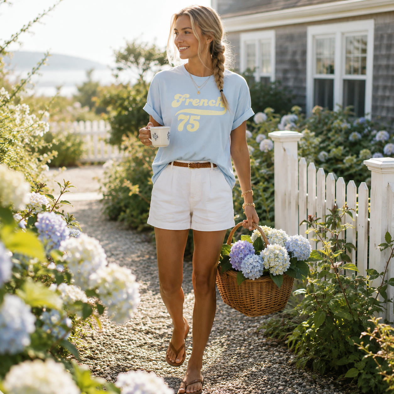 Woman walking in a garden holding a cup and a basket of flowers, wearing a light blue t-shirt with text and white shorts.